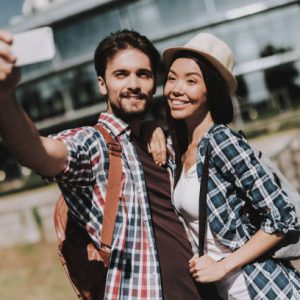 istockphoto-1061230366-612×612 Young Happy Couple with Backpacks Taking Selfie. Handsome Man and Beautiful Woman Taking Selfie in front of Glass Building by Using White Smartphone. Young Travelers Enjoying Summer Vacation